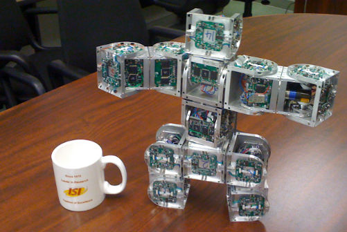 Human-shaped robot made out of cubes stretching arms next to an ISI coffee cup standing on wooden table