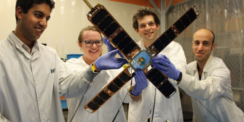 Group of four researchers holding a deployed microsatellite wearing lab coats and gloves