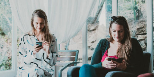 Two women sitting next to each other looking at their phones with window panes in the background and a rocky landscape