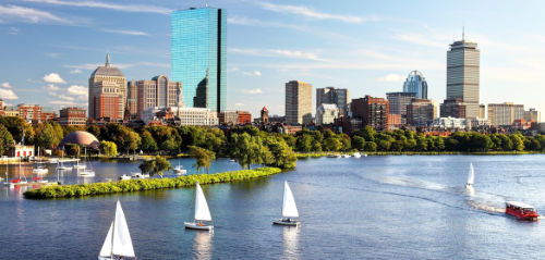 Landsacpe of a city with lake, boats, trees, and a jetty in the foreground, and a city with some tall skyscrapers and other building in the background with a clear blue sky