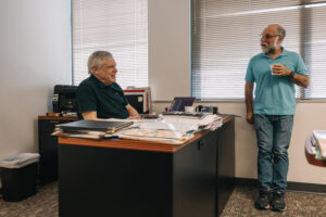 Person seated on office chair engaged in discussion with person standing across office desk